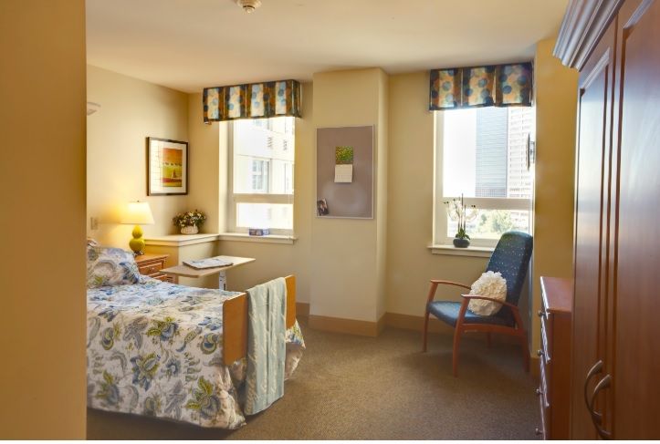 A cozy hospital room with a single bed, chair, and two windows providing natural light.