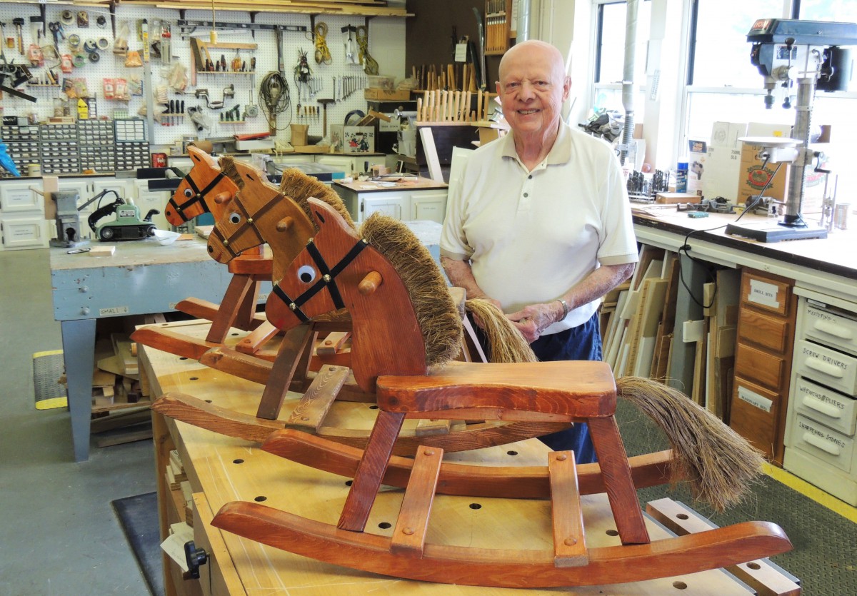 A skilled craftsman proudly displays handcrafted wooden rocking horses in a workshop filled with tools and materials.
