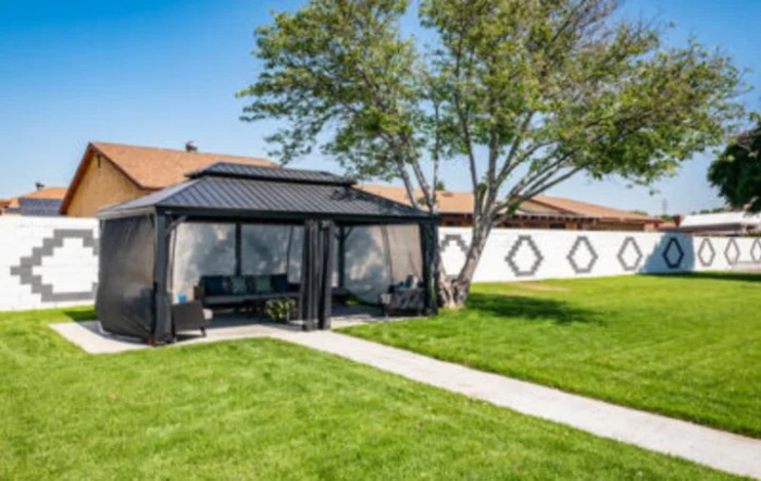 A backyard featuring a black gazebo with outdoor seating, surrounded by a lush green lawn and trees.