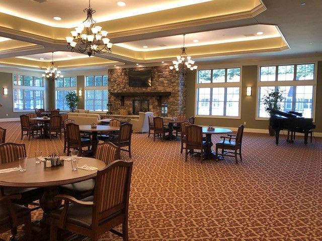 A spacious dining room with elegant decor, featuring multiple tables and chairs, ambient lighting from chandeliers, a cozy fireplace, and large windows allowing natural light.