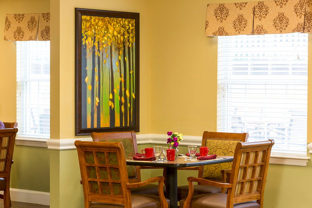 A cozy dining area with a round table, decorated with red cups and small flowers, surrounded by wooden chairs. A vibrant painting of trees adorns the wall.