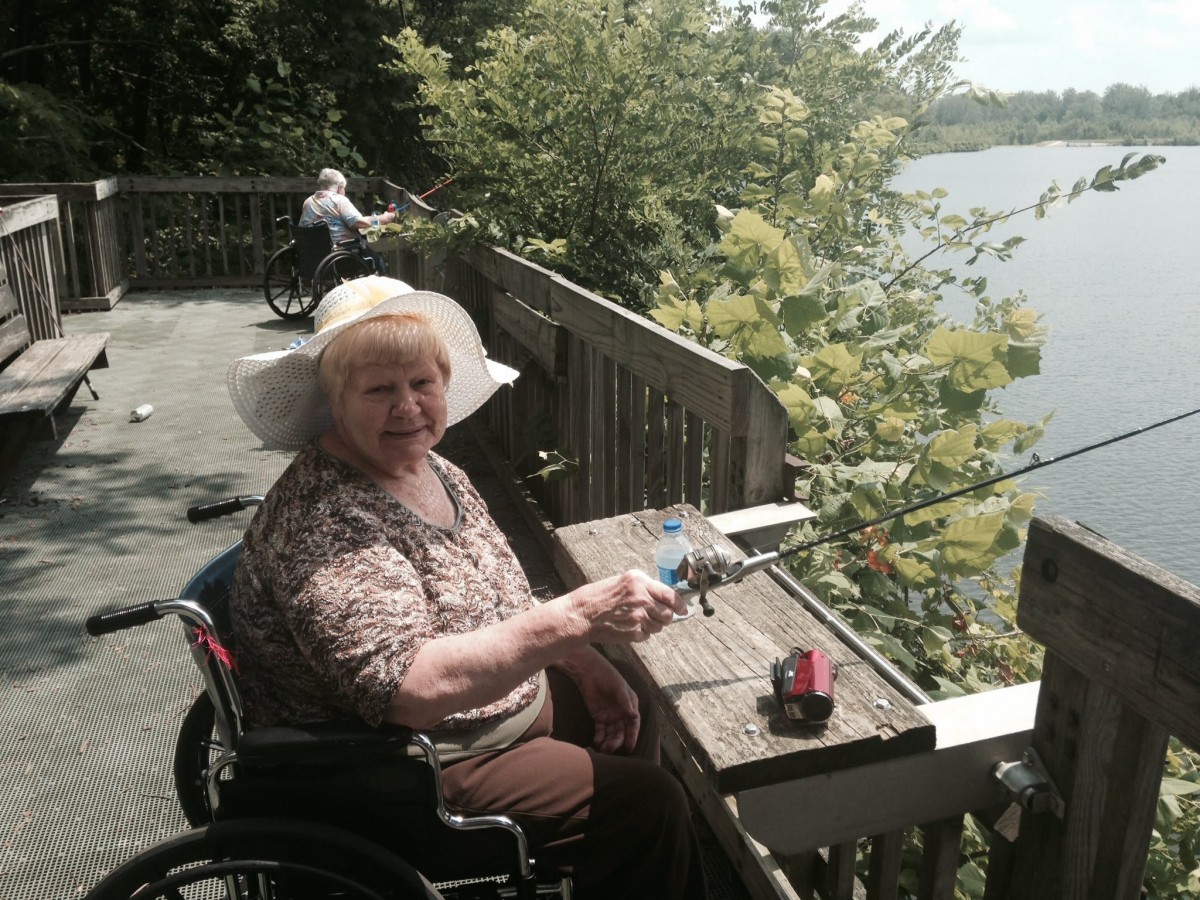 An elderly woman in a wheelchair enjoys a peaceful moment on a wooden deck by the lake, surrounded by lush greenery.