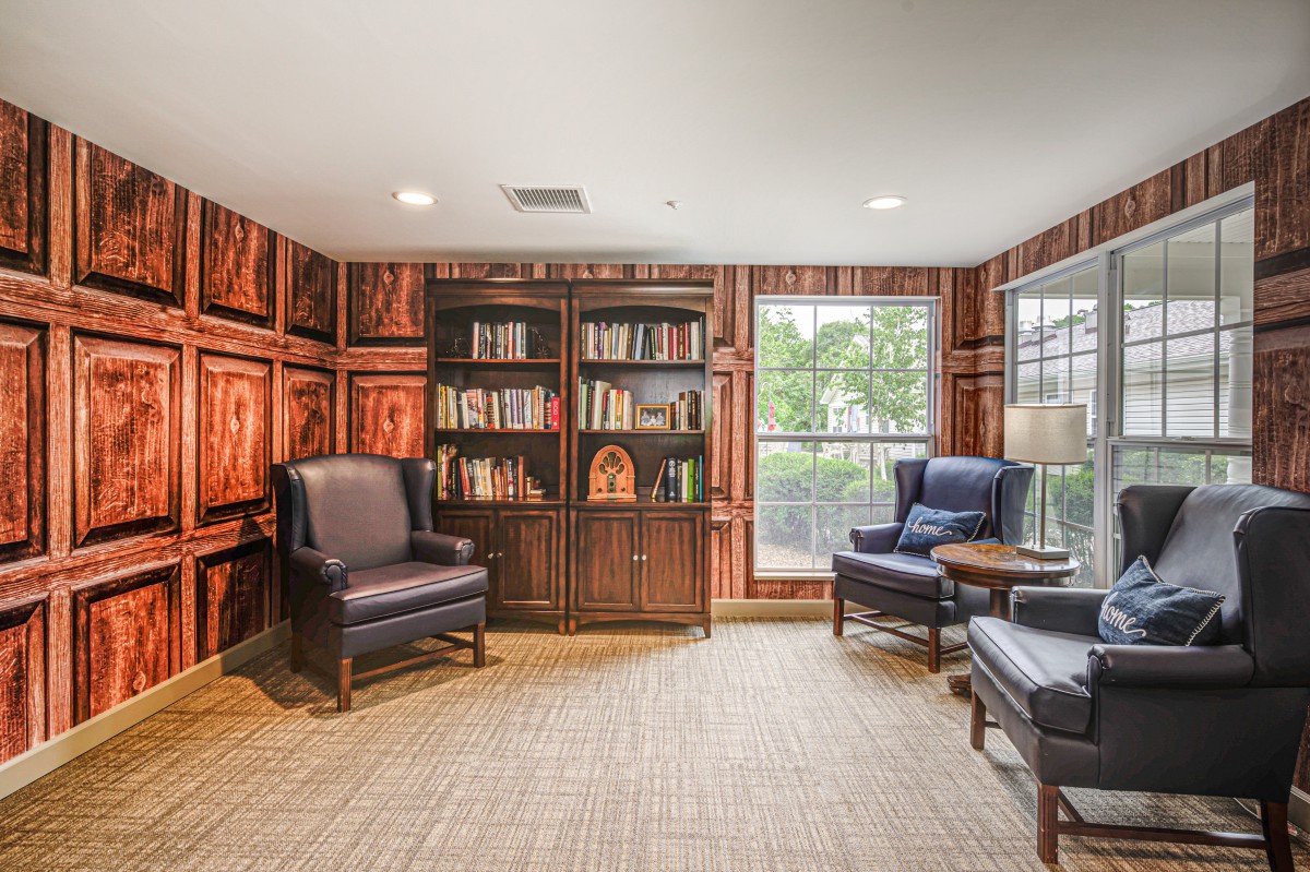 A cozy library with wooden paneling, featuring a bookshelf filled with books and comfortable armchairs for reading.