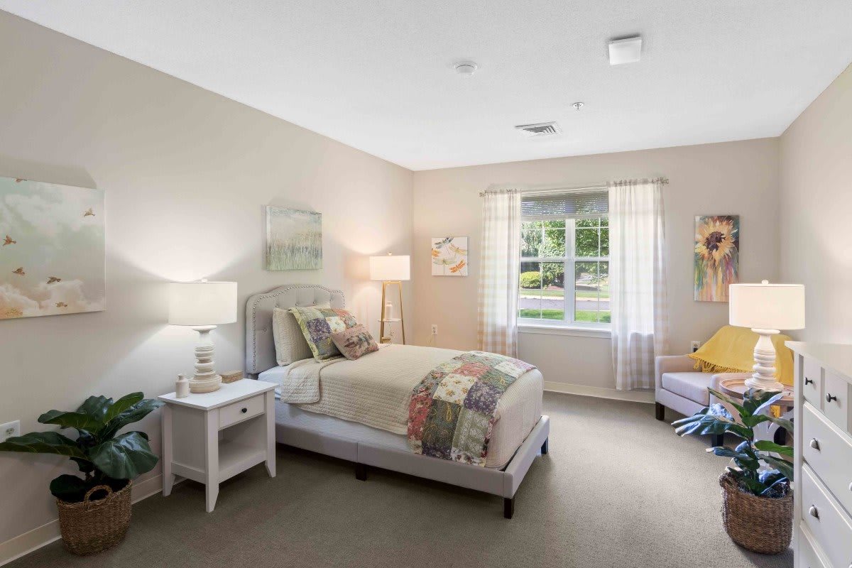 A bright and cozy bedroom with neutral tones, featuring a bed, nightstand with a lamp, and a window letting in natural light.