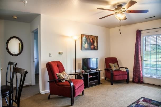 A cozy living room with red chairs, a television, artwork on the wall, and a ceiling fan.