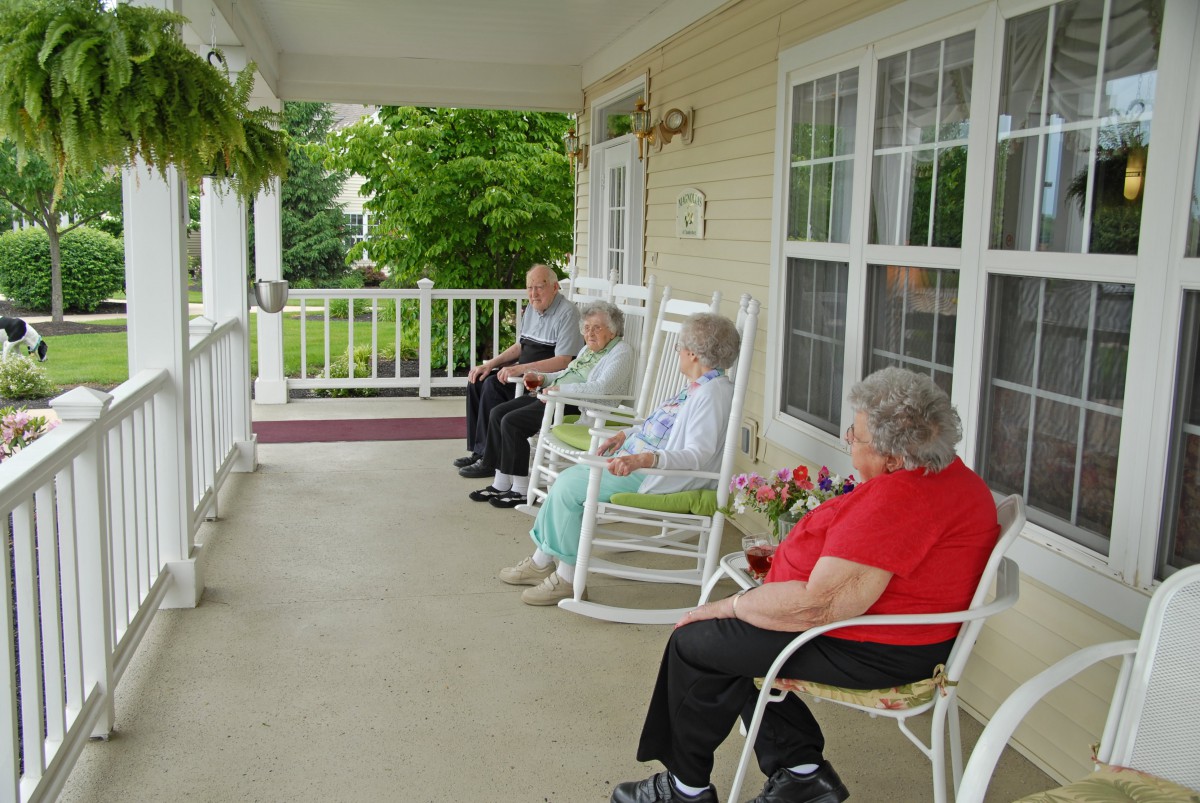 Elderly individuals relaxing in rocking chairs on a porch with a garden view.