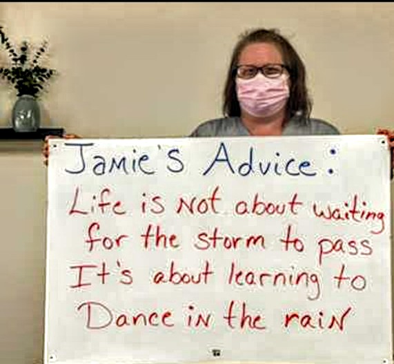 A woman wearing a mask holds a sign with an inspirational message about life and resilience.