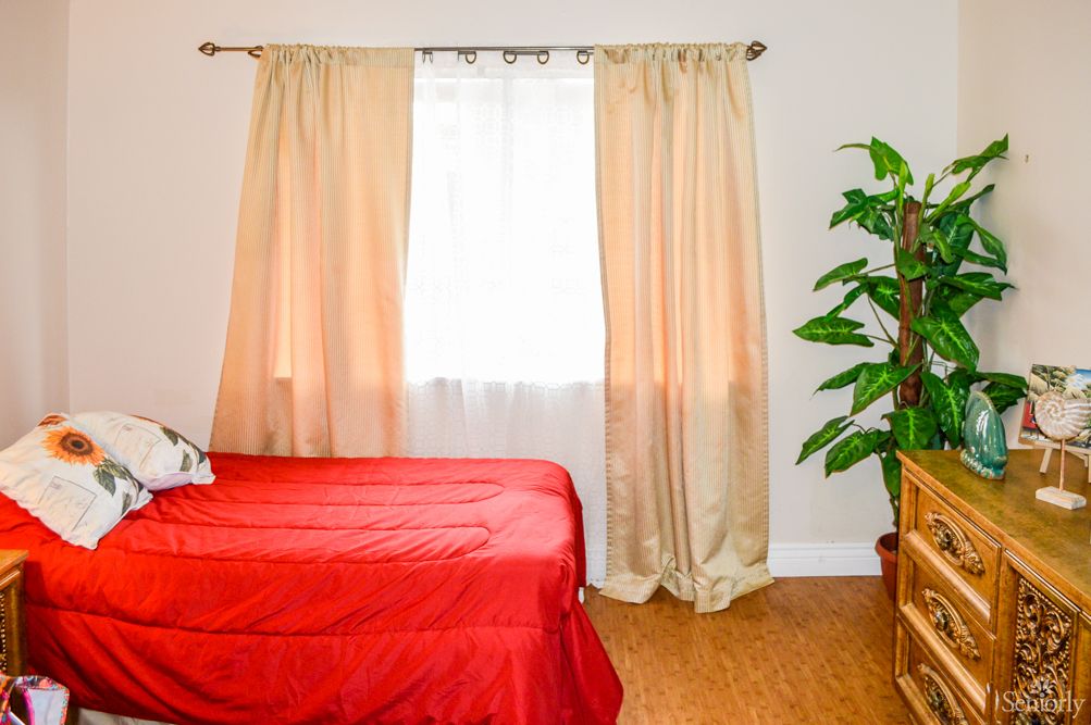 A cozy bedroom with a red bedspread, beige curtains, and a potted plant by the window.