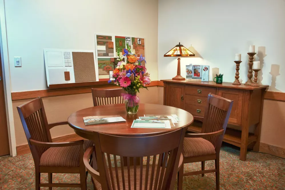 A cozy office space with a round wooden table, surrounded by chairs and adorned with a vase of colorful flowers. The room is warmly lit by a decorative lamp, and brochures are neatly arranged on a nearby counter.