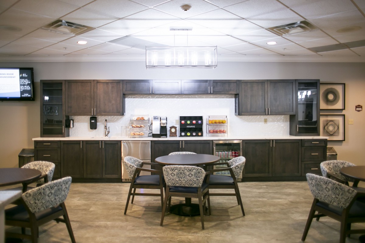 A modern office break room with a central table, chairs, and kitchen amenities.