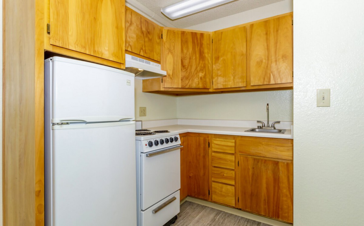 A compact apartment kitchen with wooden cabinets, a refrigerator, stove, and sink.