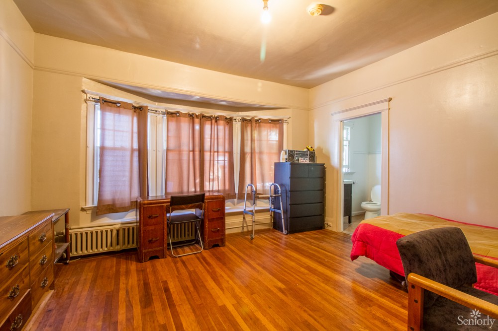 A cozy bedroom with hardwood floors, featuring a bed, desk, dresser, and large windows with curtains.