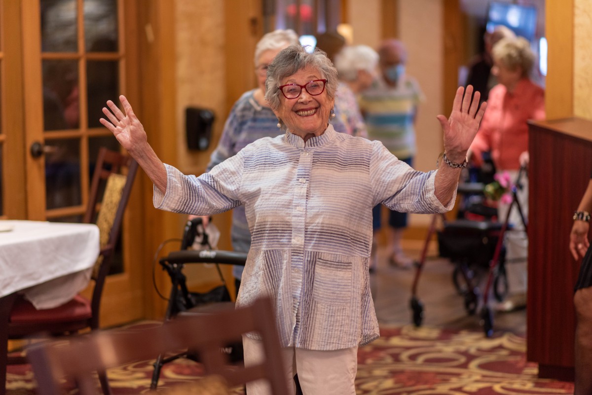 An elderly woman joyfully raises her arms in a community setting.