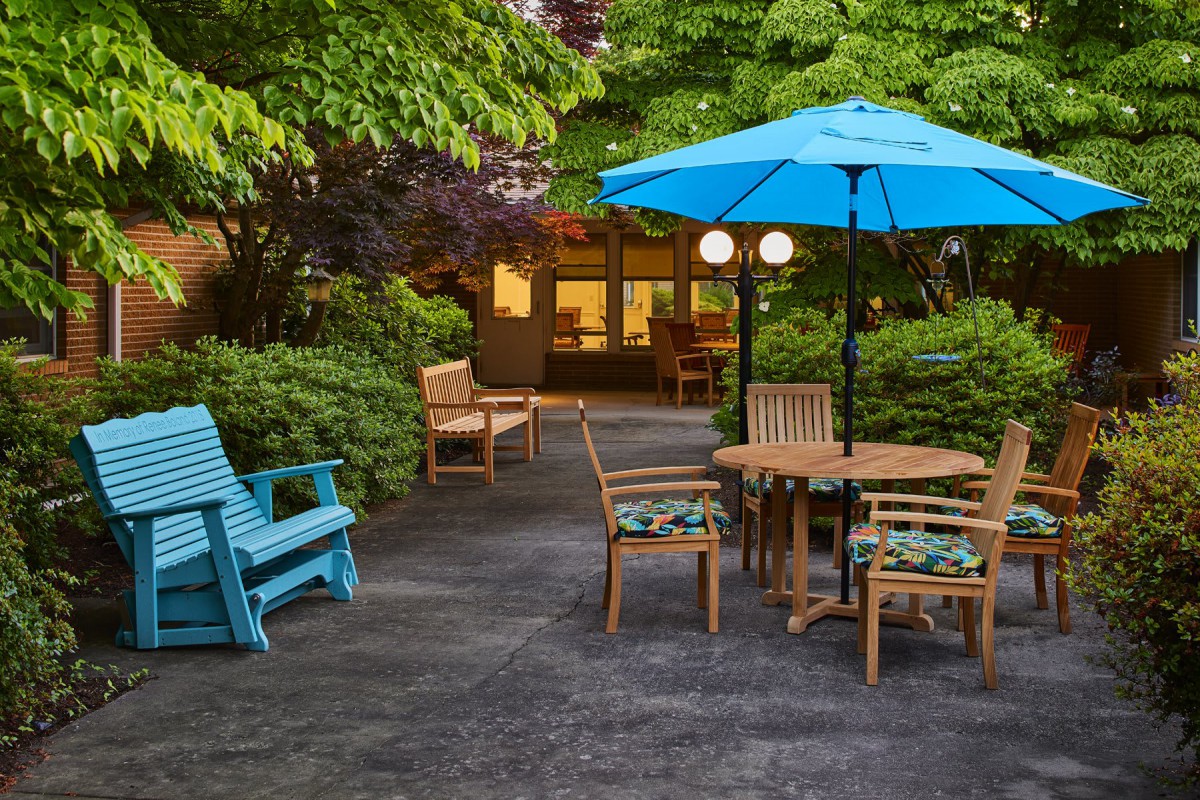 A cozy patio featuring wooden furniture, a blue umbrella, and lush greenery.