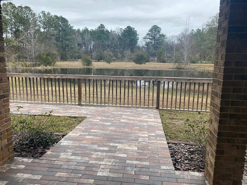 A view of a serene lake from a brick patio, surrounded by trees and fenced with wooden rails.