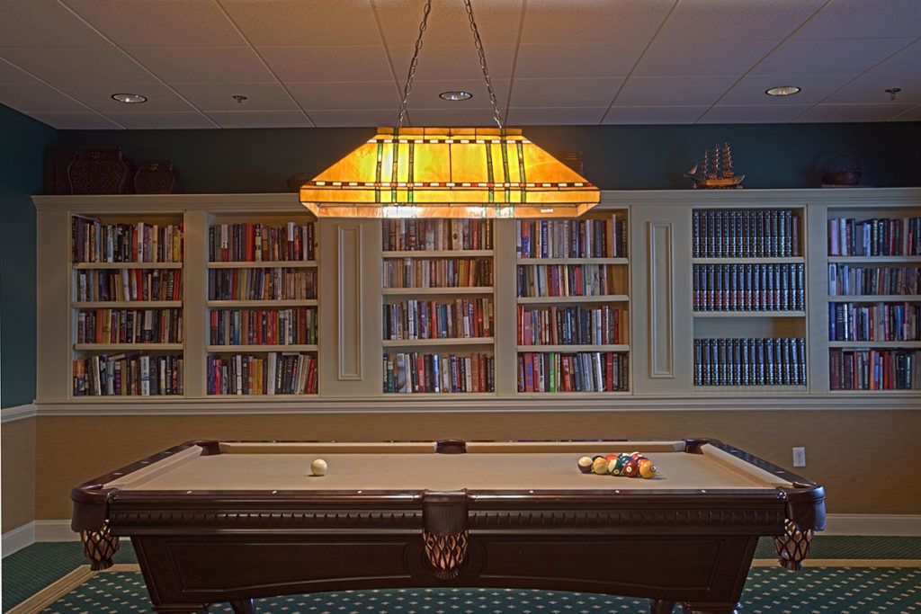 A cozy room featuring a billiard table and bookshelves, illuminated by a warm hanging lamp.