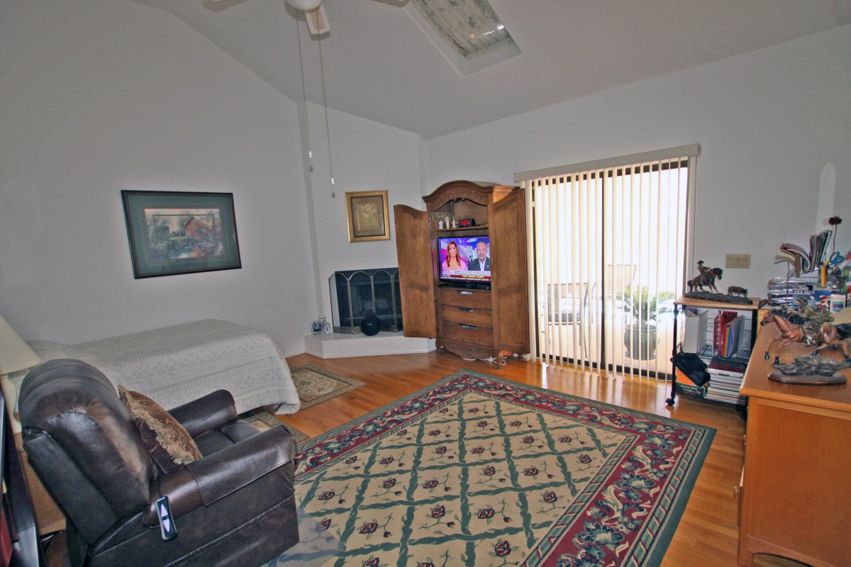A cozy living room with a comfortable armchair, a patterned rug, and a television inside a wooden cabinet. Natural light streams in through the sliding glass door.