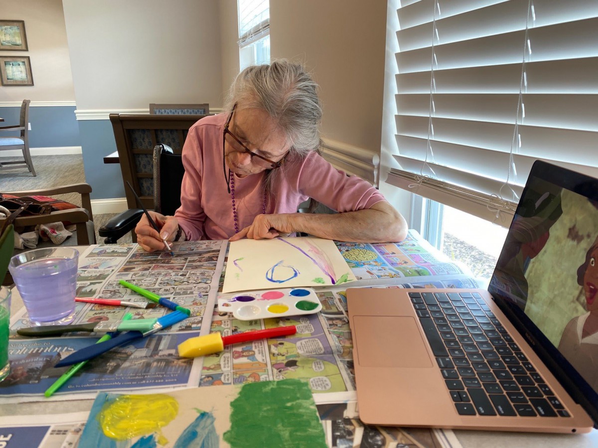 An older adult engaged in painting at a table, surrounded by art supplies and using a laptop for reference.