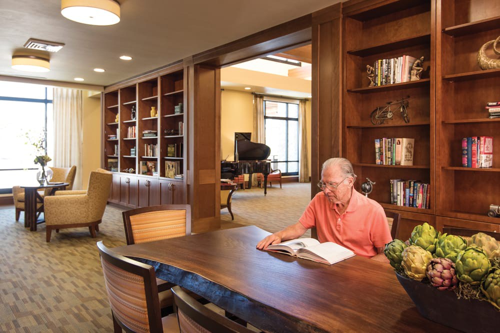 A man reads a book at a table in a cozy library setting, surrounded by bookshelves.