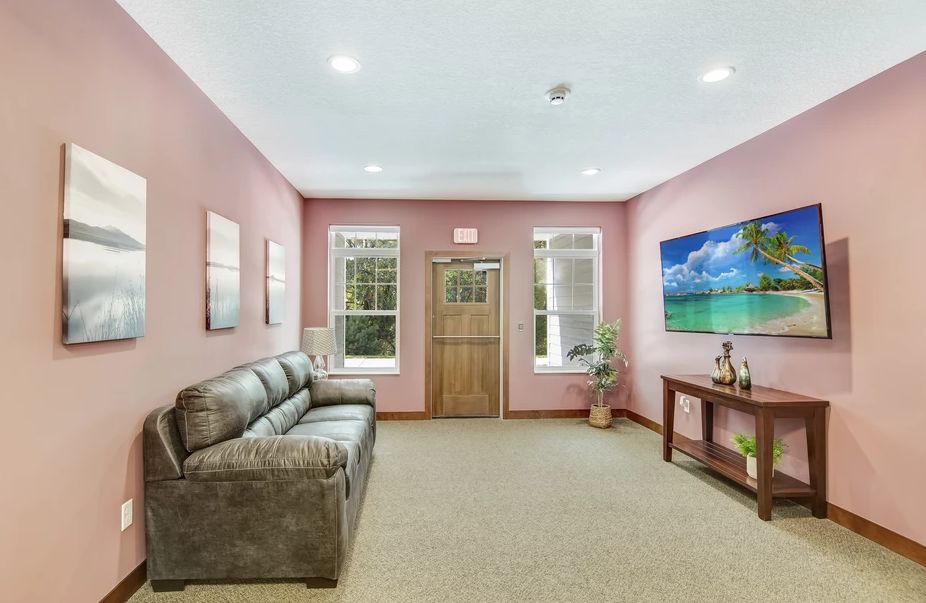 A cozy living room with pink walls, featuring a comfortable sofa, wall art, and natural lighting from large windows.