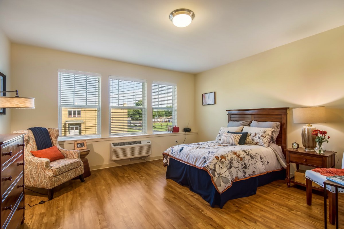 A cozy bedroom with a wooden floor, featuring a comfortable bed and ample natural light from large windows.