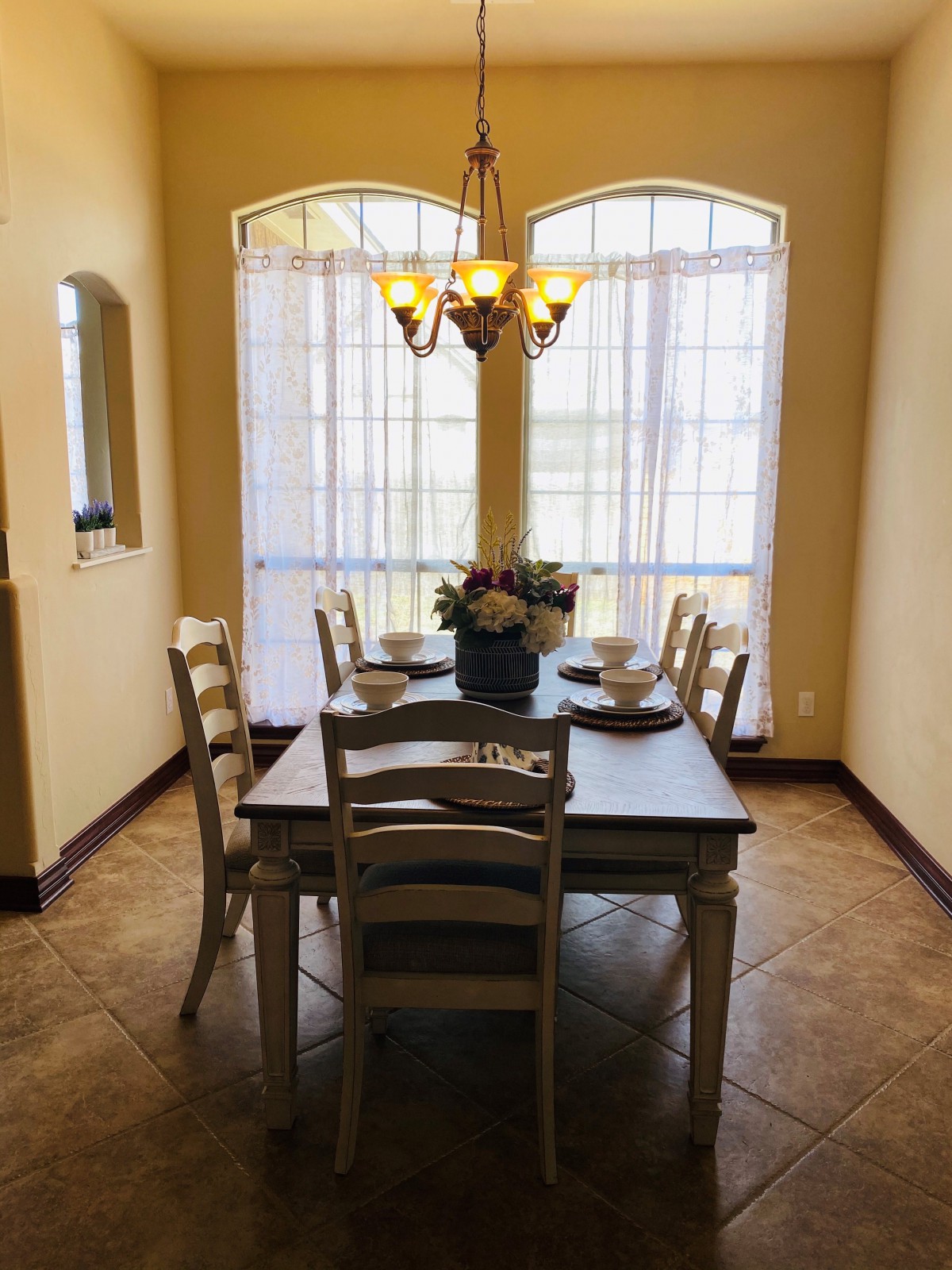 A cozy dining room with a wooden table set for four, illuminated by a chandelier and natural light from large windows.