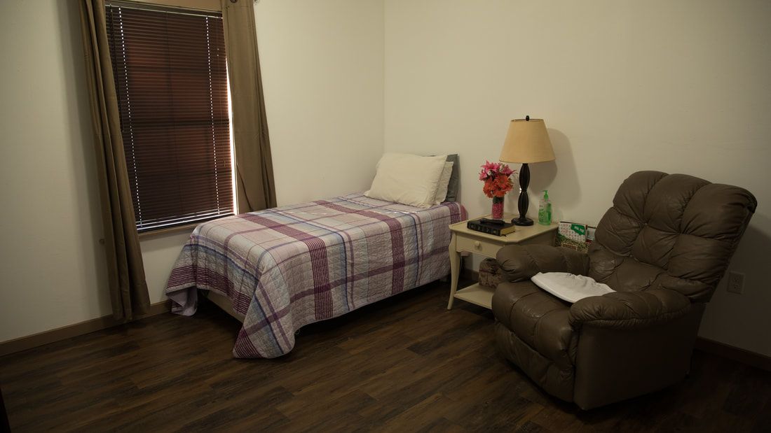 A cozy bedroom with a single bed, armchair, and side table adorned with a lamp and flowers.