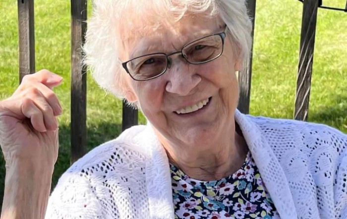 An elderly woman with glasses smiles warmly while sitting outdoors on a sunny day.