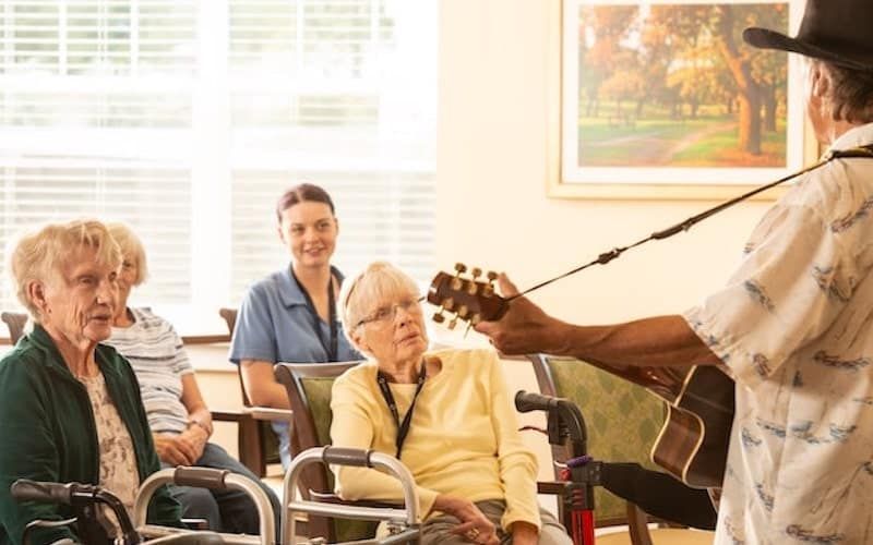 A musician plays guitar for an attentive audience in a nursing home setting.