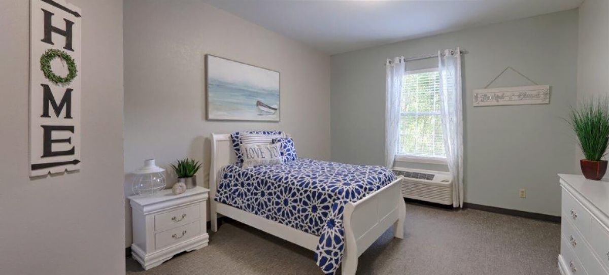 A cozy bedroom featuring a single bed with blue patterned bedding, white furniture, and natural light streaming through a window.
