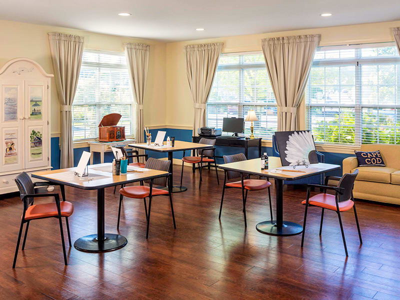 A well-lit study area with tables and chairs, featuring large windows with curtains and a wood floor.
