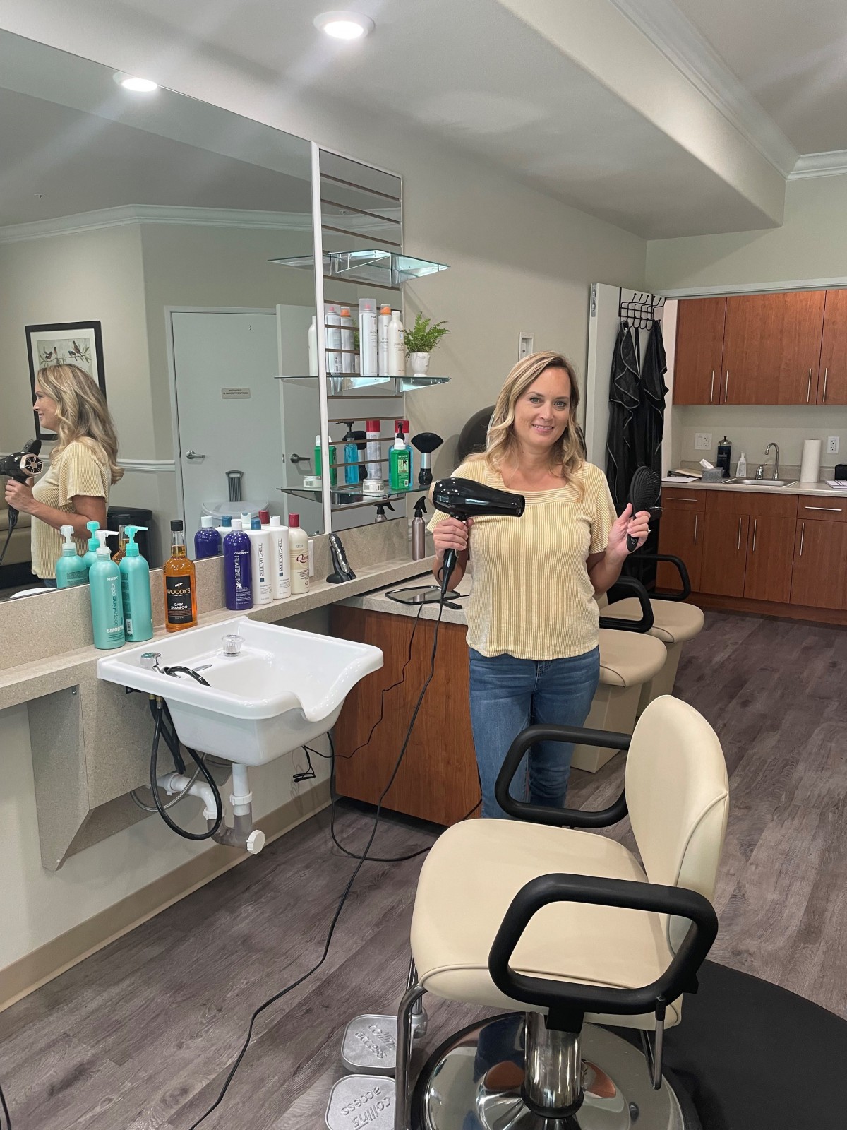 A woman stands in a hair salon holding a blow dryer, surrounded by hair care products.