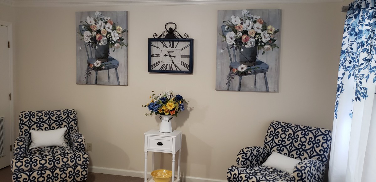A cozy interior scene featuring floral wall art, a decorative clock, and a vase of flowers on a white side table.