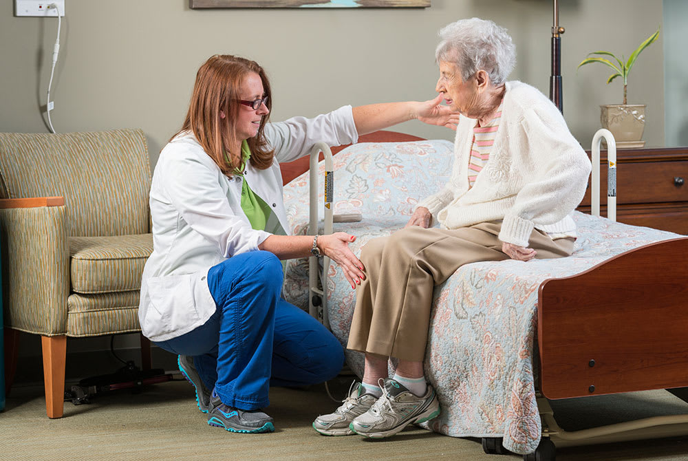 A healthcare worker kneels to interact with an elderly woman sitting on a bed.