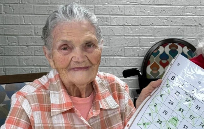 An elderly woman smiling while holding a bingo card indoors.