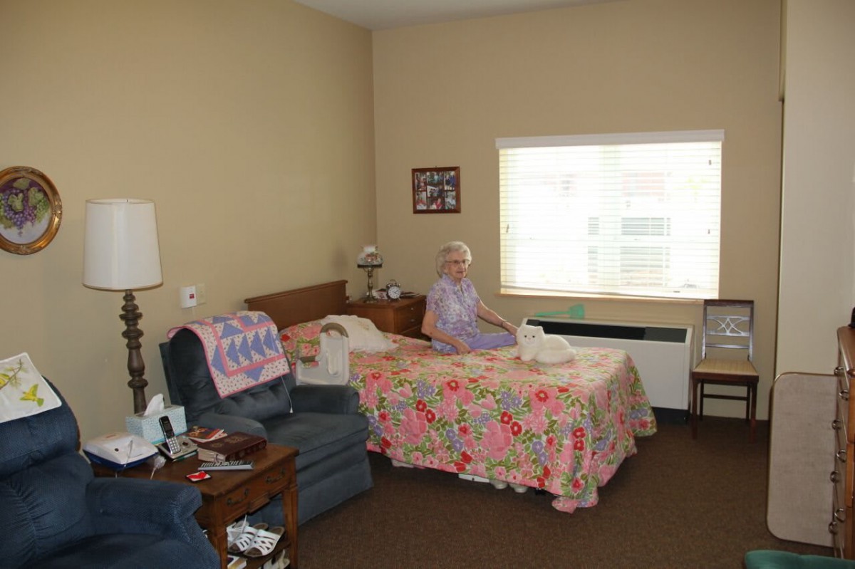 An elderly person sitting on a bed in a nursing home room with cozy furnishings.