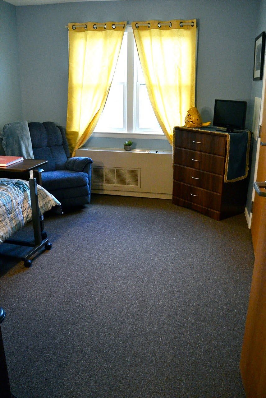 A cozy dorm room with a bed, chair, and dresser near a window with yellow curtains.