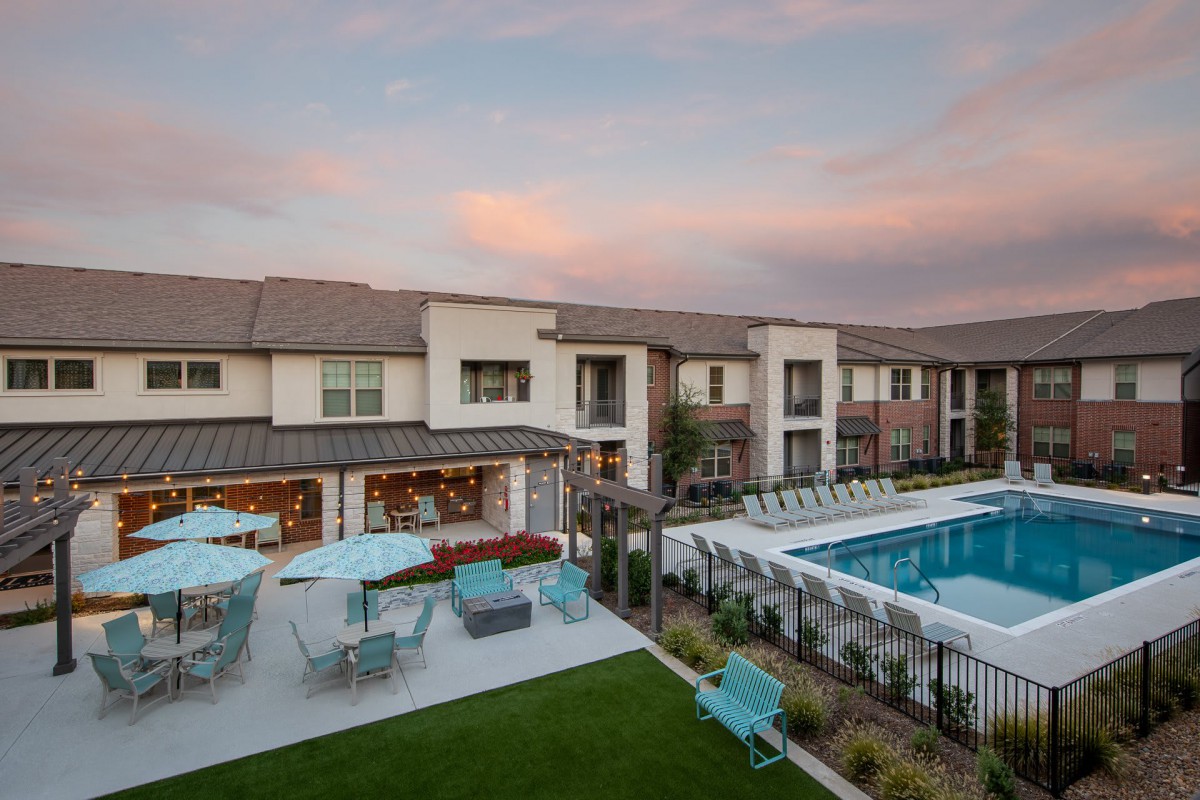 A modern residential complex featuring a large swimming pool and outdoor seating area under a beautiful sunset sky.