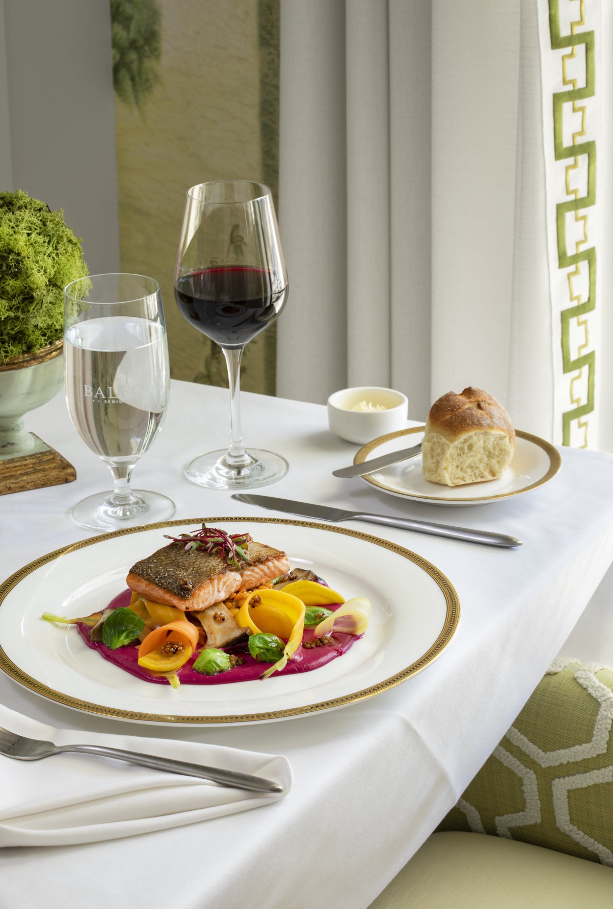 A beautifully set table with a gourmet salmon dish, a glass of red wine, and bread roll in an elegant dining setting.