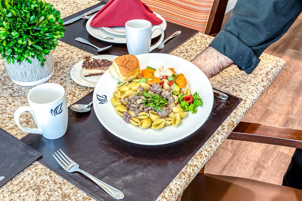 A dining setup featuring a plate of pasta with vegetables, a bread roll, and coffee on a table.