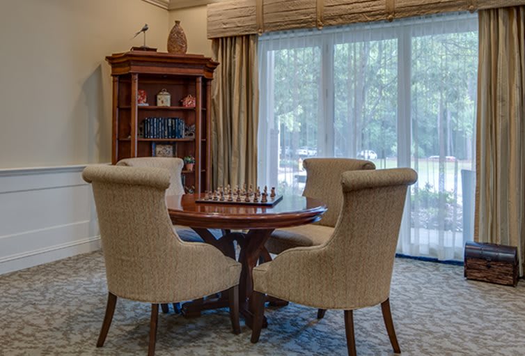 A cozy dining room with a chess set on the table, surrounded by upholstered chairs and a bookshelf in the corner.