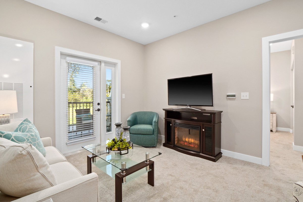 A modern living room featuring a fireplace with a television above it, a glass coffee table, and seating area with a sofa and armchair.