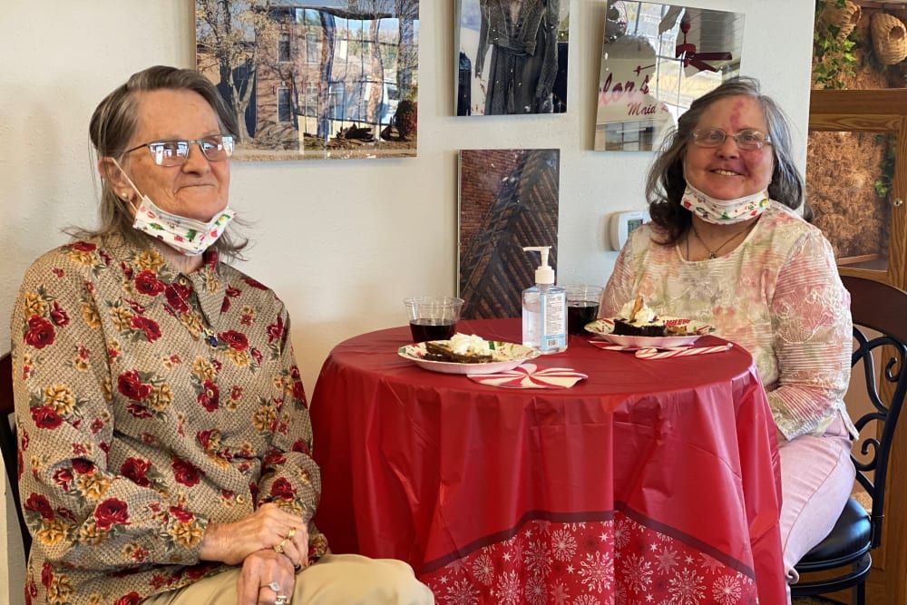 Two seniors sitting at a small table with red tablecloth, enjoying snacks and drinks indoors.