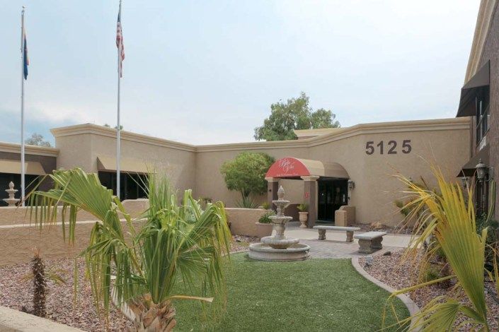 Exterior view of a building entrance with a flag, fountain, and landscaped greenery.