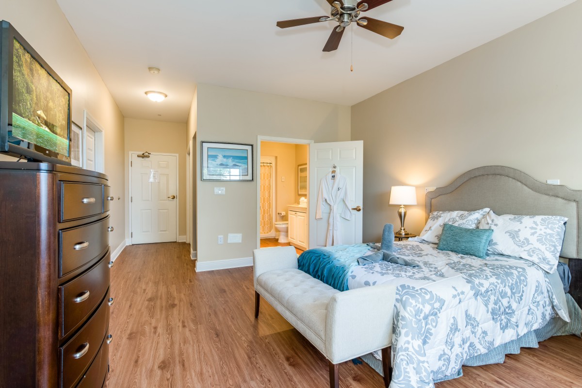 A cozy bedroom with a bed, lamp, and ceiling fan, featuring hardwood floors and light-colored decor.