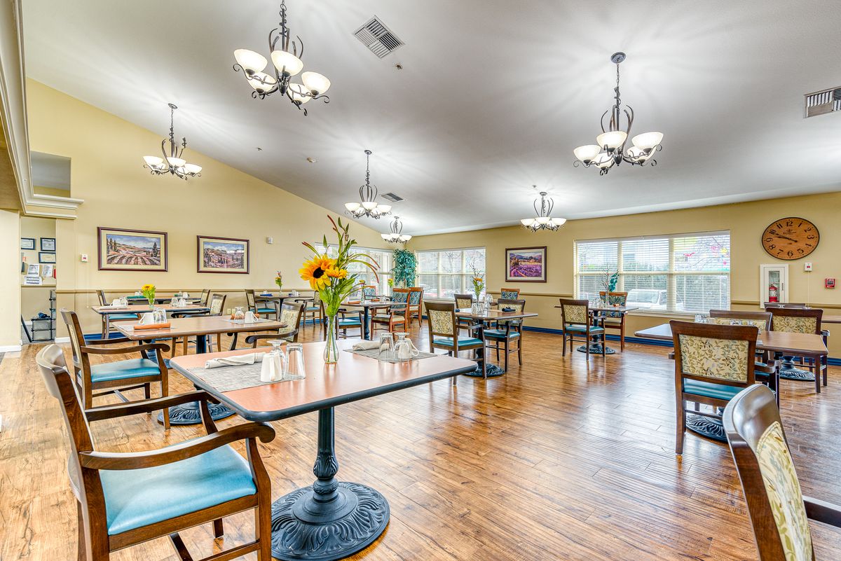 A well-lit dining area with arranged tables and chairs, decorated with flowers and paintings.