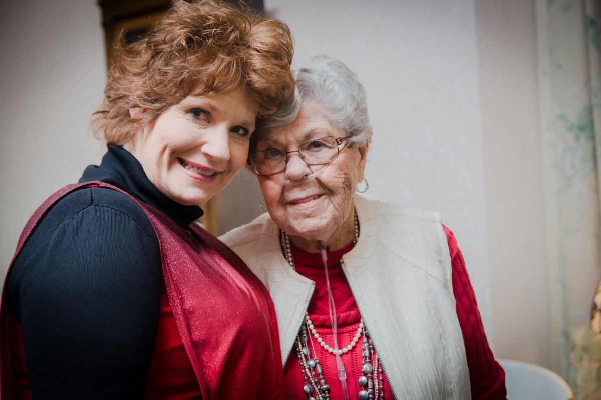 An elderly woman and a younger woman smiling together indoors.