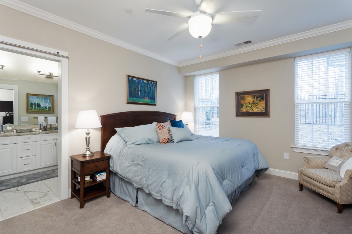 A cozy and well-lit bedroom featuring a large bed with blue bedding, flanked by nightstands with lamps, a ceiling fan, and paintings on the walls.