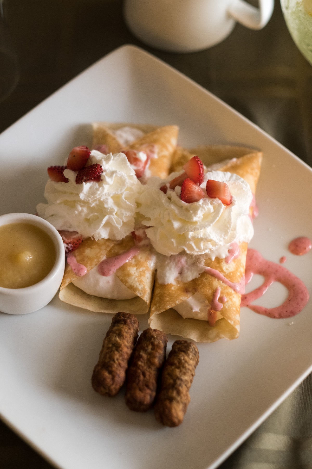 A delicious plate with strawberry crepes topped with whipped cream and served alongside breakfast sausages.