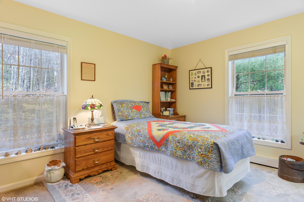 A cozy bedroom with a single bed featuring a colorful quilt, wooden nightstand and bookshelf, illuminated by natural light from two windows.